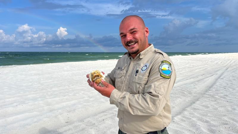 A park ranger stands on the beach holding a replica sea turtle skull. There is a rainbow in the background over the Gulf of Mexico.